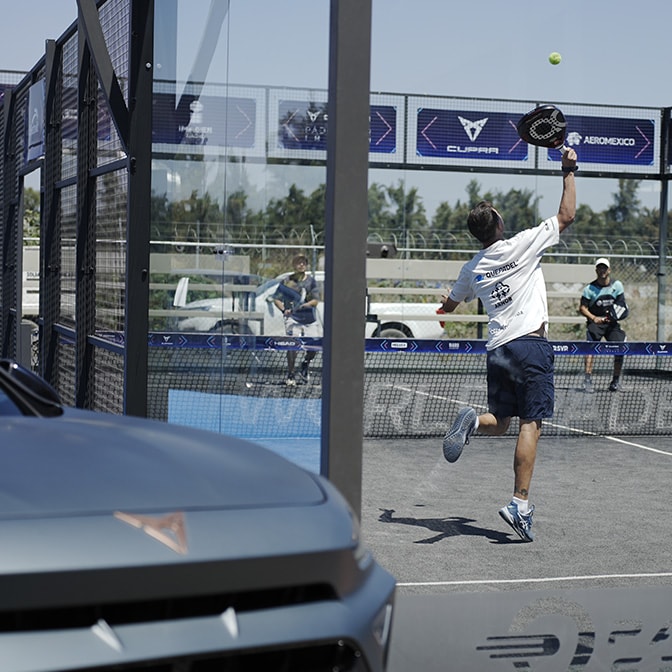 A wide-angle landscape shot at dusk showing a woman in a trench coat standing on a winding coastal road next to a CUPRA Terramar, with rocky hills and a calm sea in the background.