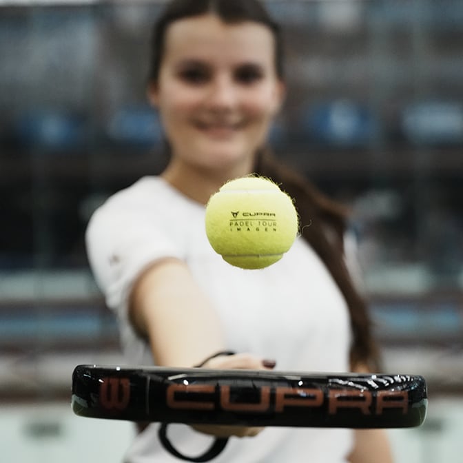 A woman leaning against a grey CUPRA Leon and looking at her phone while the car is plugged into a wall-mounted charger outside a modern, minimalist wood and concrete house.