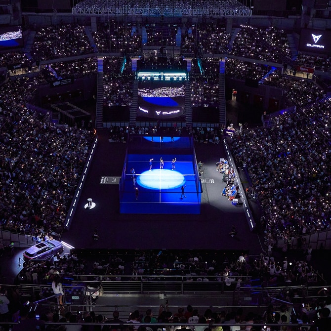 A large indoor stadium filled with spectators watching a padel match on a brightly lit court surrounded by glass walls. The audience is seated in tiers, and the court is the main focus under strong lighting.
